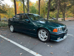 1997 BMW M3 E36 in green with aftermarket modifications parked in a scenic area during autumn.