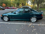 1997 BMW E36 M3 sedan in green with aftermarket wheels parked in a leafy area.