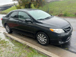 2012 Toyota Corolla LE in black parked on the street with rain-soaked surface, showcasing its sleek design.