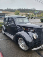 Classic 1937 Ford sedan with a partially opened hood parked on a rainy day, showcasing its vintage design.