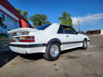 1986 Ford Mustang Foxbody with a white exterior and rear spoiler, parked in front of a mural.